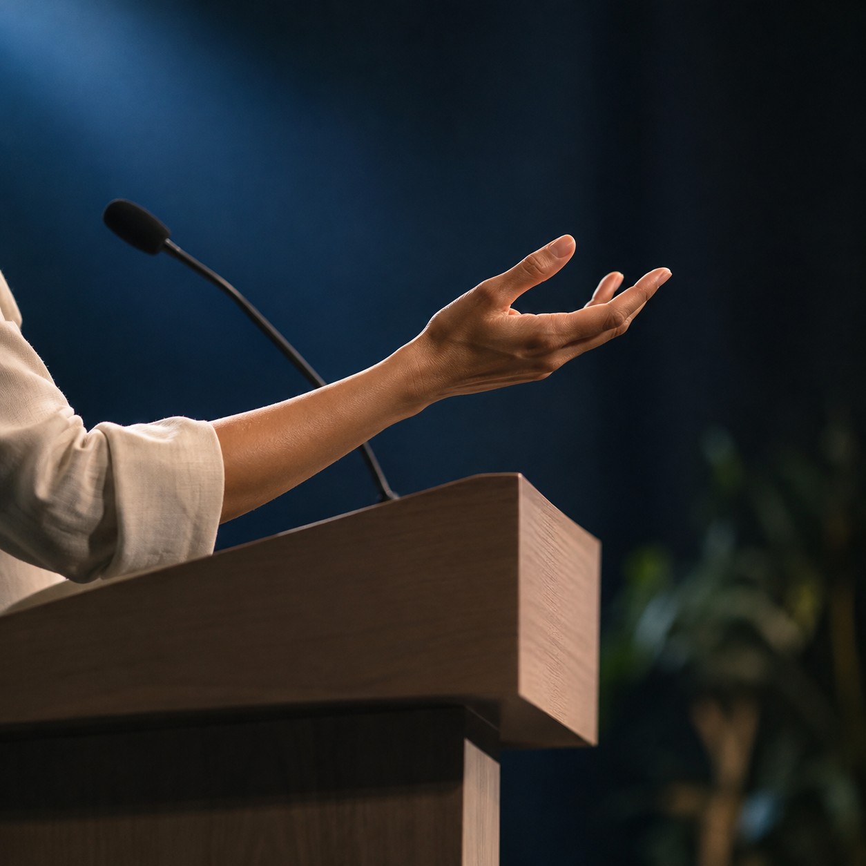 Illustrative scene: A close side view of an adult palm extended in an expressive gesture above a plain stage lectern, hand and forearm only, dark background with soft blue spotlight.