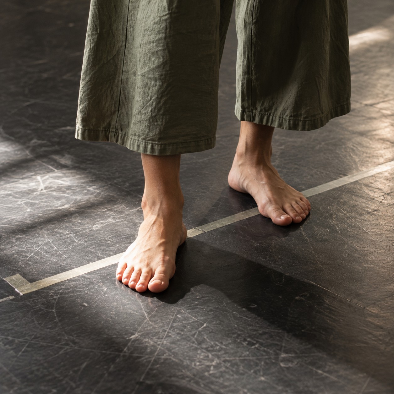 Illustrative scene: An adult planting bare feet firmly on a black rehearsal floor beside a strip of stage tape, only calves and feet, muted olive wide-leg trouser hems, overhead light.